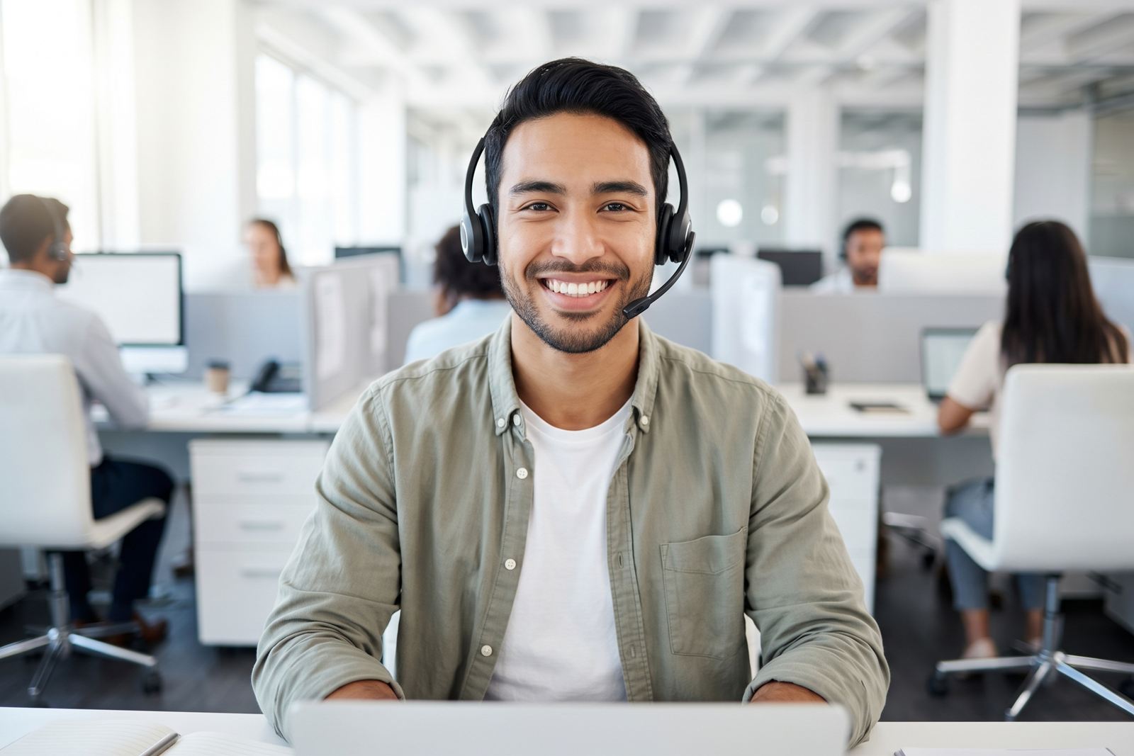 موظف خدمة عملاء يبتسم وهو يرتدي سماعة رأس، ويجلس أمام حاسوبه في مكتب مفتوح.