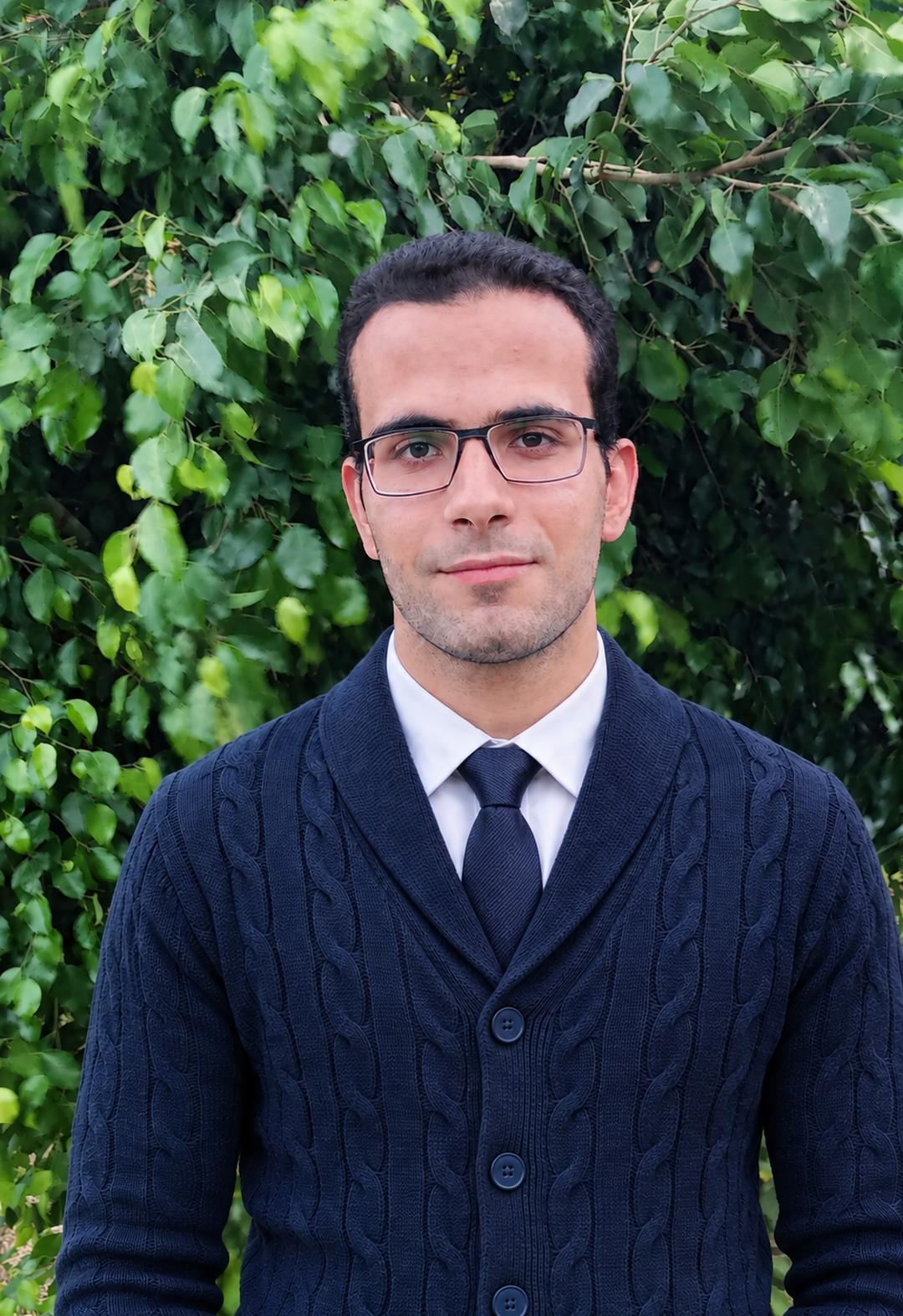 عمر محمد، مدرب اللغة الإنجليزية للكورس، يبتسم أمام خلفية نباتية.
